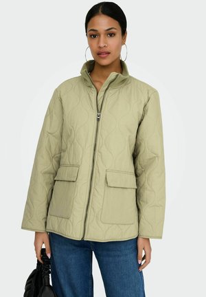 Femme portant une veste matelassée vert olive clair avec poches avant et fermeture éclair, assortie à un jean bleu et de grandes boucles d'oreilles créoles, tenant un sac noir.