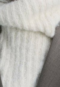 Valkoinen ribattua neuletta oleva kaulus harmaan bleiserin alla, jossa on pehmeä, pörröinen rakenne ja näkyvä ommel reunan päällä.