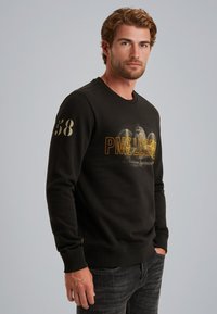 Sudadera negra con el texto "PME LEGEND" en dorado, con cuello redondo y puños de canalé. El modelo la lleva puesta con jeans oscuros.