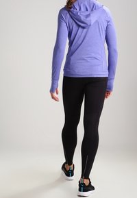 Hoodie sportivo lungo a maniche viola con cappuccio e aperture per i pollici, abbinato a leggings neri con cerniere alle caviglie e un design textured.