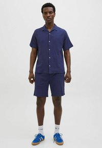Chemise à manches courtes bleu marine avec fermeture à boutons, assortie à un short. Le tissu semble texturé. Les chaussures sont bleues avec des semelles beiges.