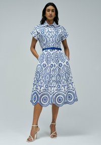 Vestido blanco con bordado floral azul, mangas cortas, cuello tipo polo, cintura entallada con cinturón a juego y dobladillo con ondas.