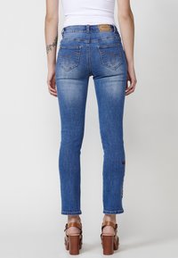 Sittande blå denimjeans med en blekt textur, som har broderade lappar på benen och bakfickorna, samt en midja med medelhöjd.