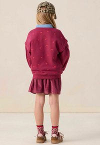 Enfant portant un sweat à capuche bordeaux avec de petits cœurs rouges, une jupe assortie, une casquette à imprimé léopard et des chaussures, de dos contre un fond uni.