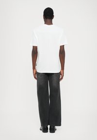 Hvit bomulls T-skjorte med korte ermer, kombinert med vide, falmede sorte jeans og sorte sko, sett fra baksiden.