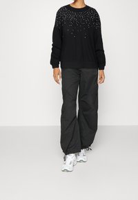 Persona vistiendo un suéter negro de manga larga con un patrón de puntos blancos, pantalones cargo negros holgados y zapatillas blancas sobre un fondo liso.