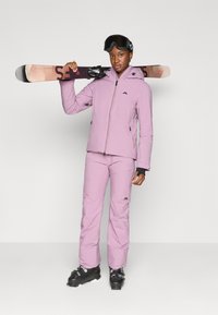 Roze skijack met bijpassende broek, voorzien van een ritssluiting aan de voorkant, logo-accenten en zwarte skischoenen. Het model houdt skies vast en draagt een zwarte helm.