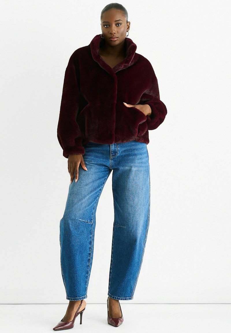 Woman wearing a dark burgundy faux fur jacket, blue high-waisted jeans, and burgundy high-heeled slingback shoes, standing against white background.
