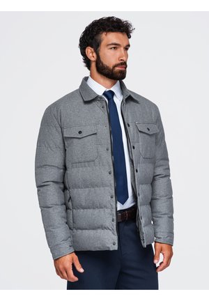 Homme aux cheveux bouclés et à la barbe portant une veste matelassée grise sur une chemise blanche et une cravate bleue sur un fond uni.
