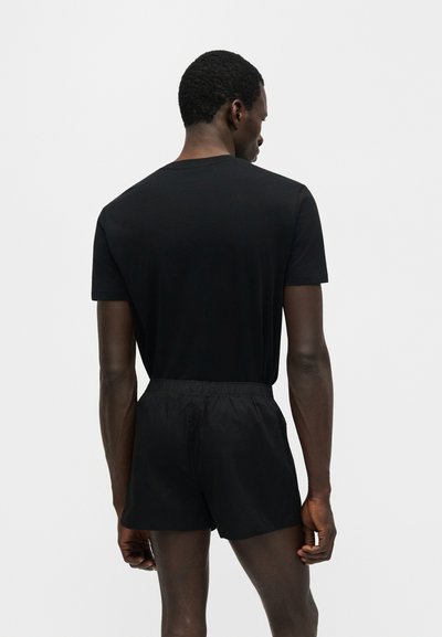 Homme portant un t-shirt noir à manches courtes et un short noir avec taille élastique, debout de dos face à la caméra sur fond blanc.