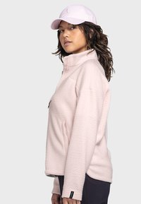 Helle rosa Fleecejacke mit hohem Kragen, durchgehendem Reißverschluss und seitlichen Taschen. Kombiniert mit einer passenden rosa Kappe mit einem Logo.