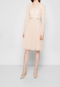 Abito rosa pallido con maniche lunghe in pizzo, scollatura a V profonda e una gonna morbida e plissettata. Presenta dettagli in perline e una cintura in satin in vita.