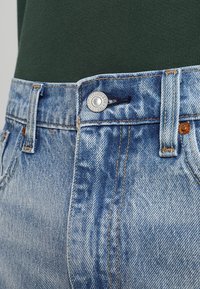Jeans de mezclilla azul claro con una cintura alta, cierre de botón de metal y costuras en marrón contrastante. La tela presenta un aspecto desgastado y texturizado.
