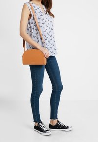 Ljusblå blus med fjärilsprint, mörkblå smaljeans och en liten orange axelväska; svarta sneakers med vita sulor och snörning.