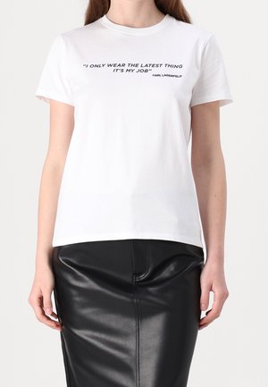 Femme portant un t-shirt blanc avec un texte noir « Je ne porte que les dernières tendances, c’est mon métier » et une jupe en cuir noire.