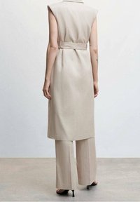 Beżowa sukienka bez rękawów z paskiem w talii, noszona na spodniach z szerokimi nogawkami w tym samym kolorze. Gładka tkanina o minimalistycznym wzornictwie, bez widocznych wzorów.
