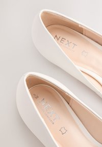 Hvide flade sko med glat læderoverflade, rundet tå og blødt beige indvendigt. Har sølvfarvet branding på indersålen.