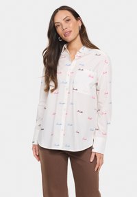 Camicia bianca con bottoni, decorata con la scritta "Smile" ripetuta in blu, rosa e nero. Presenta una tasca sul petto e maniche lunghe. Tessuto di cotone liscio.