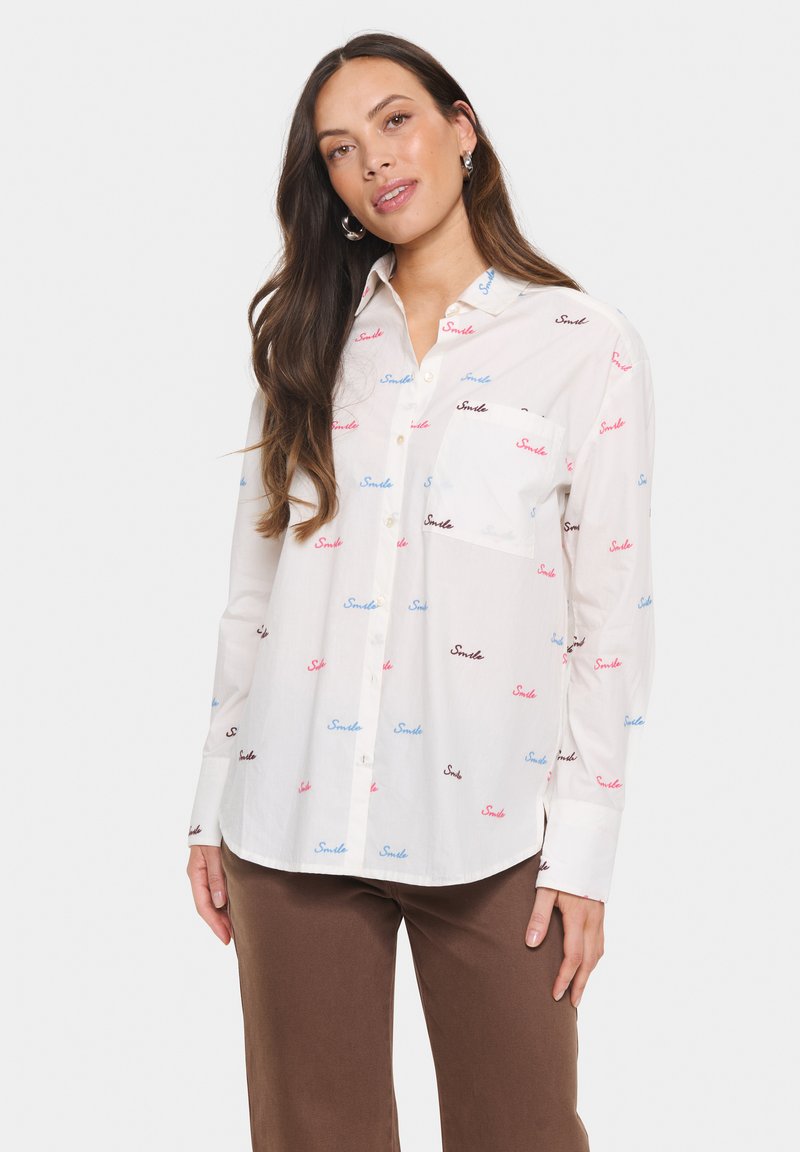 Camicia bianca con bottoni, decorata con la scritta "Smile" ripetuta in blu, rosa e nero. Presenta una tasca sul petto e maniche lunghe. Tessuto di cotone liscio.