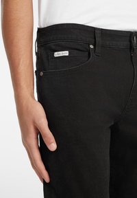 Svart denimjeans med en tettsittende design. Har en logoetikett på frontlommen og metallknappdetaljer. Glatt tekstur og klassisk snitt.