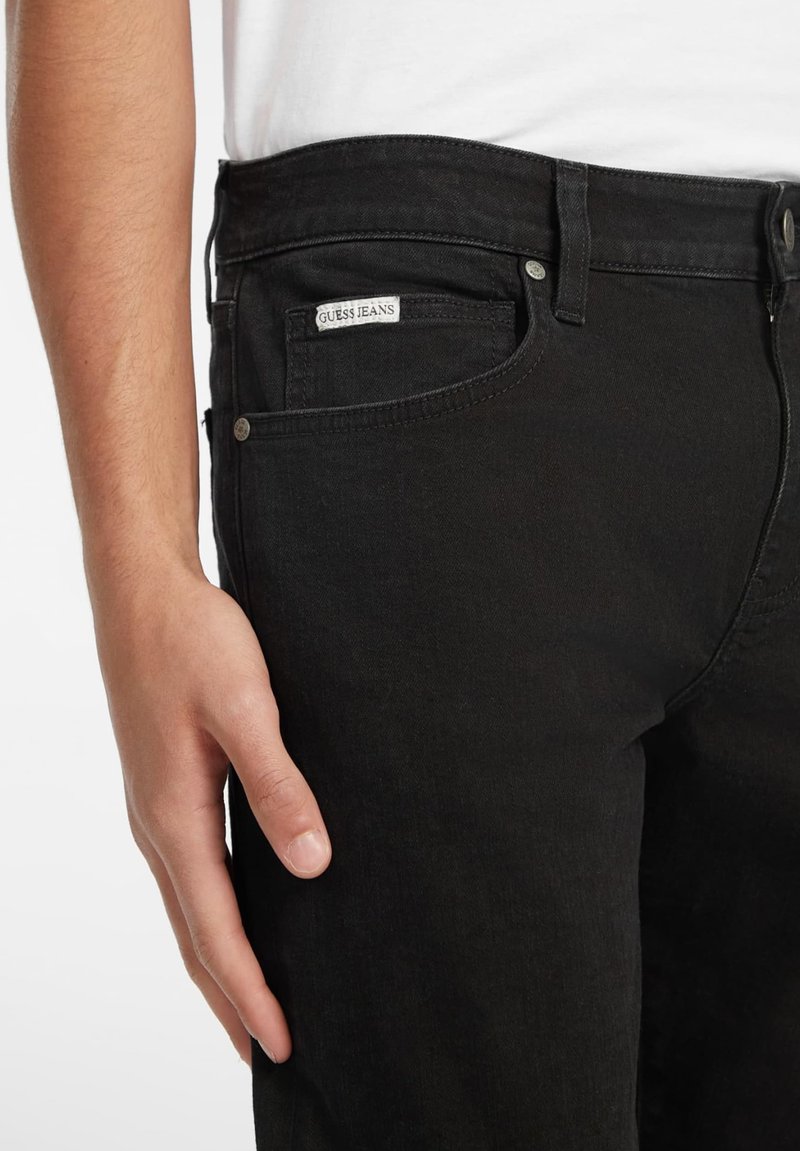 Jean en denim noir avec une coupe ajustée. Présente une étiquette logo sur la poche avant et des accents en métal sur les boutons. Texture lisse et coupe classique.