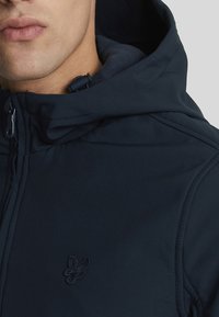 Giacca con cappuccio blu navy con chiusura a zip, foderata internamente in modo morbido e con logo ricamato sul petto. Presenta cuciture strutturate e cappuccio regolabile.
