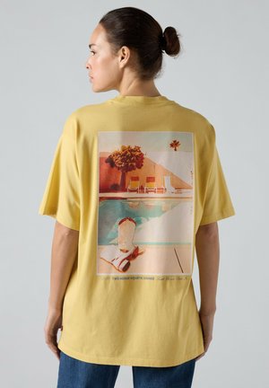 Kobieta z włosami związanymi w koka, ubrana w żółtą koszulkę z grafiką w stylu retro przedstawiającą leżaki przy basenie, drzewo i stopy osoby z tyłu.