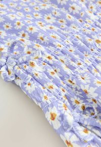 Blomstermønstret stof i lavendel med hvide kamilleblomster og gule midter. Har en gathered tekstur med en lille sløjfedetalje.