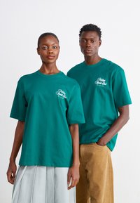 Teal bomulds t-shirts med korte ærmer, med et lille hvidt logo på brystet. Den mandlige model har brune corduroy bukser på.