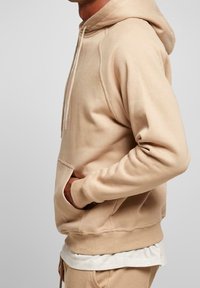 Beige katoenen hoodie met een trekkoord, buidelzak en geribbelde zoom. Zachte textuur, ontspannen pasvorm en naadloos schouderontwerp.