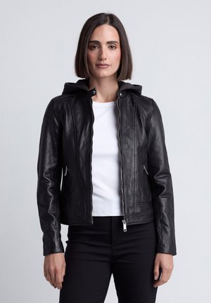 Vrouw met steil donker haar, een zwarte leren jas over een wit overhemd en zwarte broek dragend, staand tegen een effen lichte achtergrond.