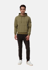 Olive green hoodie with a front pocket, adjustable drawstring, and ribbed cuffs, paired with dark grey pants and white sneakers.