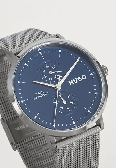 Montre-bracelet à bracelet en maille argentée avec cadran bleu foncé, aiguilles blanches, sous-cadrans jour et date, étiquetée "HUGO" et résistante à l'eau jusqu'à 30 mètres.