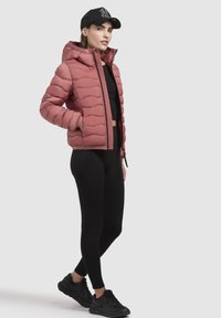 Padded pink jacket with a hood, featuring a quilted design and front zipper. Worn over a black crop top and black leggings with black sneakers.