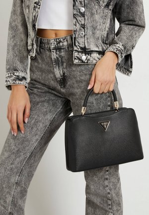 Kobieta w kurtce i spodniach z denimu acid-wash, trzymająca czarną torebkę Guess z fakturowaną powierzchnią i złotymi okuciami przy uchwycie.