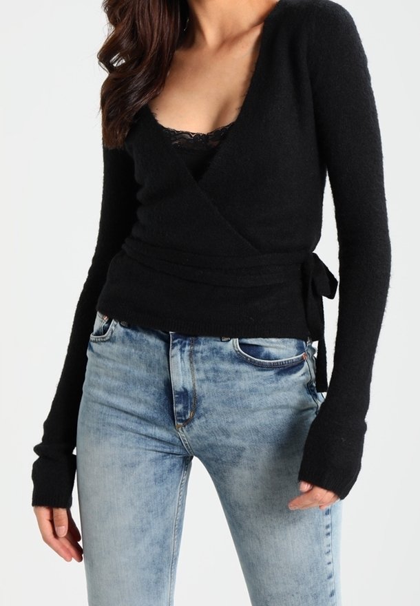 Femme portant un pull cache-cœur noir par-dessus une camisole en dentelle noire avec un jean en denim bleu clair, debout devant un fond uni.