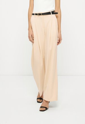 Femme portant un pantalon beige plissé à jambes larges avec une ceinture noire et des sandales à talons ouvertes noires, debout sur un fond blanc.