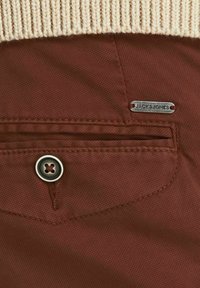 Hnědá látka s texturovaným povrchem, s stříbrnou etiketou "JACK & JONES" a zapínáním na knoflík. Pružný béžový úplet na horním okraji.