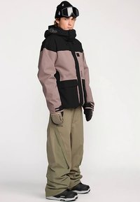 Sneeuwjas in zwart en roze met een capuchon, meerdere zakken en ritssluitingen. Gecombineerd met losse, lichtgroene sneeuwbroek en zwarte laarzen.