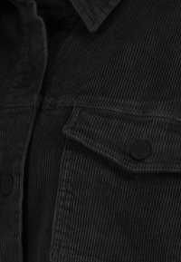 Zwarte corduroy blouse met verticale ribbels, voorzien van een gestructureerde kraag en zwarte drukknoopzakken. De zachte textuur versterkt het ontwerp.