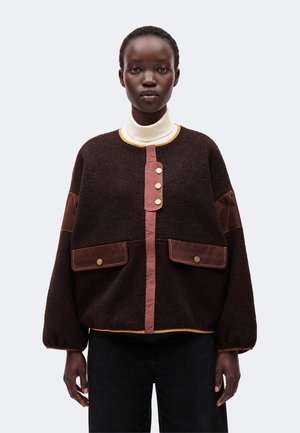 Femme portant une veste ample marron foncé avec des bordures roses et des boutons dorés, un col roulé blanc et un pantalon noir, debout devant un fond uni.