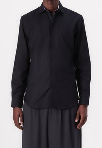 Camicia nera a maniche lunghe con colletto classico, chiusura con bottoni e una trama liscia e opaca; abbinata a pantaloni scuri e ampi.