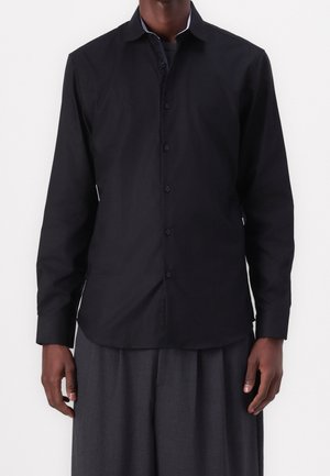 Czarna koszula z długim rękawem, klasycznym kołnierzykiem, zapięciem na guziki i gładką, matową fakturą; zestawiona z ciemnymi, luźnymi spodniami.