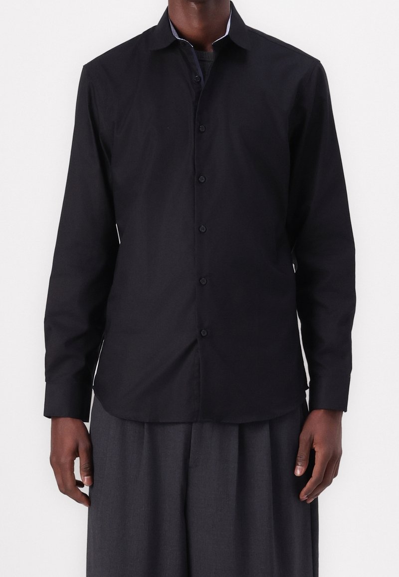 Camicia nera a maniche lunghe con colletto classico, chiusura con bottoni e una trama liscia e opaca; abbinata a pantaloni scuri e ampi.