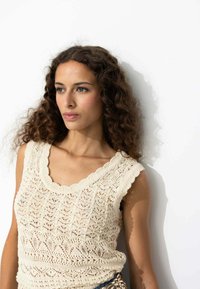 Débardeur en tricot beige avec un col à volants, présentant un motif délicat en dentelle et des bretelles larges, sur un fond blanc uni.