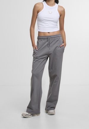 Femme portant un débardeur blanc court et un pantalon de jogging gris ample, les mains dans les poches, debout sur un fond blanc uni.