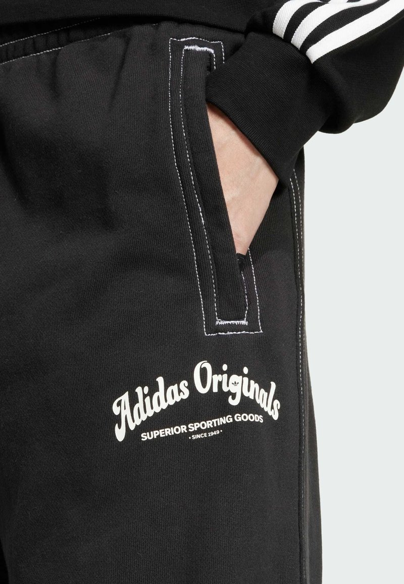Czarne dresowe spodnie z białymi szwami i kieszenią. Zawierają logo "Adidas Originals" w białym kolorze, podkreślając sportowy design i fakturę materiału.