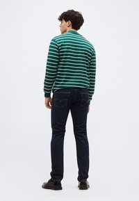 Άνδρας που φοράει πράσινο και λευκό ριγέ πουκάμισο με μακριά μανίκια, σκούρο μπλε τζιν και μαύρα παπούτσια, όρθιος με την πλάτη προς την κάμερα.