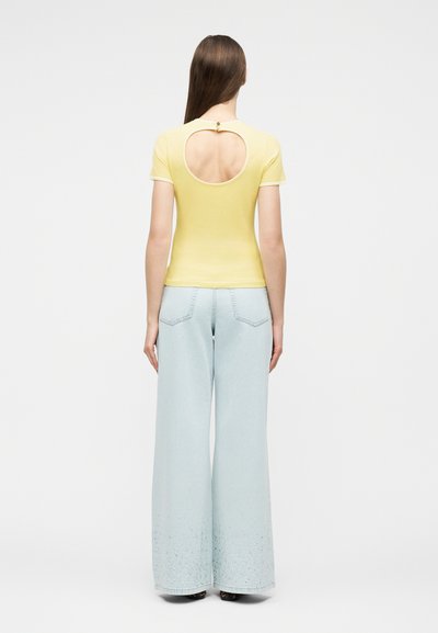 Γυναίκα με μακριά καστανά μαλλιά φοράει κίτρινο τοπ με κοντά μανίκια και στρογγυλή ανοιχτή πλάτη, μαζί με ανοιχτό μπλε τζιν παντελόνι με φαρδιά μπατζάκια.