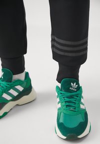 Sneakers verdi con strisce bianche, materiale texturizzato, suola in gomma e dettagli viola. Pantaloni neri con tre strisce orizzontali grigie.